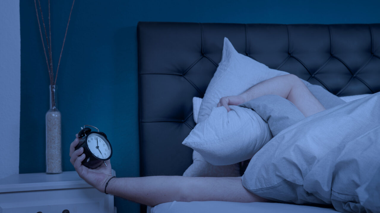 Sleepless man with pillow over face grabbing alarm clock.