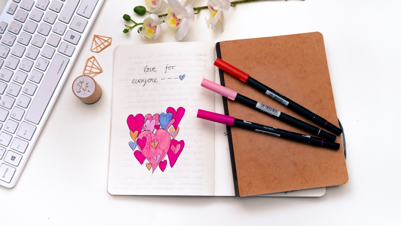 Close up of doodled hearts and the words love for everybody in a journal with fine liners, and keyboard nearby on white background.