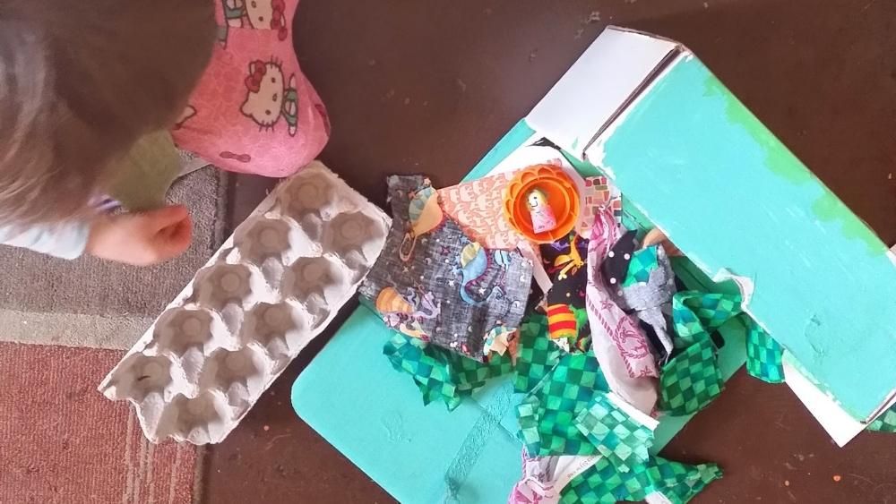 A toddler stands next to a dumped out box of fabric scraps and wooden peg dolls