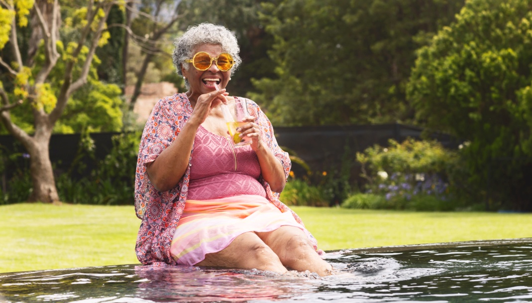 Older woman happy wearing bright sunglasses drinking water while sitting with legs in a pool.