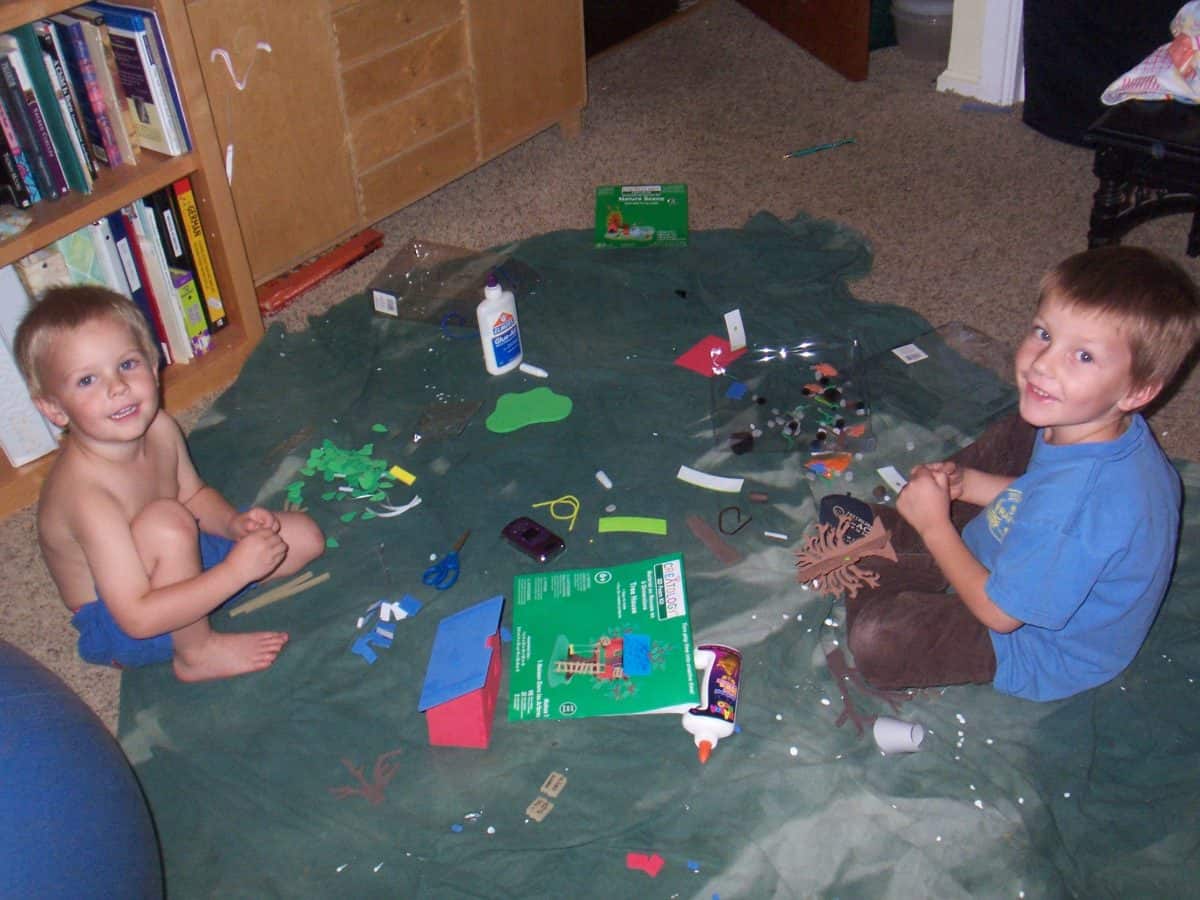 Two boys sit on a blanket with a craft mess all around.